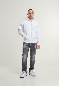 Sudadera blanca con cremallera, grandes bolsillos frontales y capucha, combinada con jeans gris oscuro y zapatillas blancas con acentos azules. Textura suave.