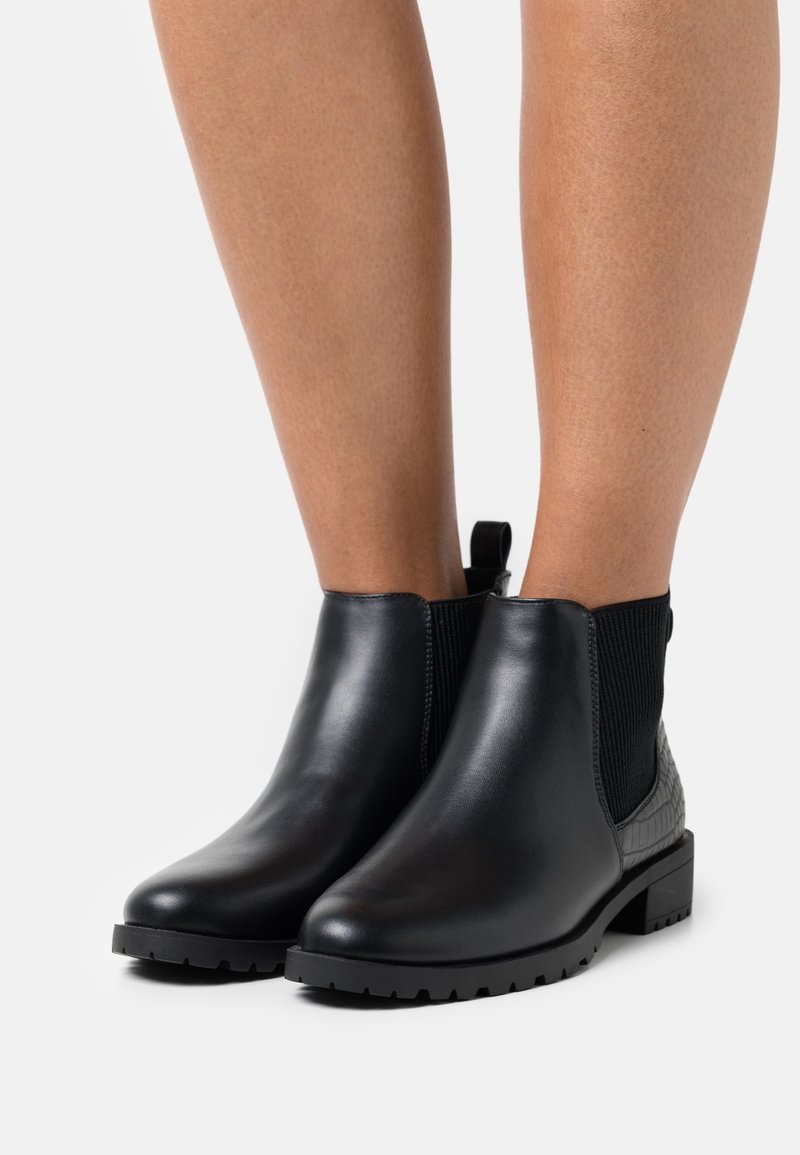 Bottines en cuir noir avec panneaux latéraux élastiques et talon texturé, portées sur des jambes nues sur fond blanc.