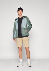 Giacca leggera a cappuccio a blocchi di colore in verde scuro e azzurro chiaro, abbinata a pantaloni cargo beige. Modello che indossa sneakers bianche.