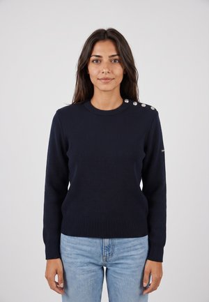 Donna con lunghi capelli scuri indossa un maglione blu navy con bottoni argento sulla spalla e jeans azzurri, in piedi davanti a uno sfondo uniforme.