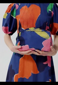 Osoba koja nosi plavu haljinu s velikim apstraktnim narančastim, ružičastim, zelenim i žutim oblicima, držeći pravokutnu torbicu s odgovarajućim uzorkom.