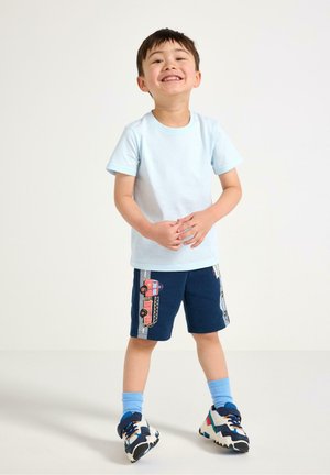 Smilende dreng iført en lyseblå t-shirt, marineblå shorts med et brandbilsdesign, blå sokker og sneakers i flere farver, stående på en ensfarvet baggrund.