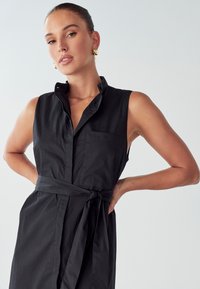 Abito nero senza maniche con collo a camicia, tasca frontale e dettaglio in vita legato, realizzato in tessuto liscio. Design minimalista.