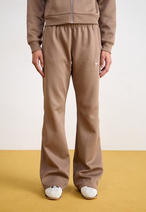 Personne portant un pantalon de jogging beige à jambes larges avec un petit logo blanc Nike sur la cuisse gauche, des baskets blanches et une veste zippée beige assortie.