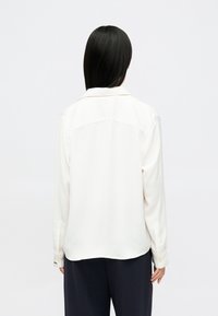 Camisa blanca de manga larga con cuello abotonado y una textura de tejido suave. Presenta un dobladillo curvado y puños abotonados.