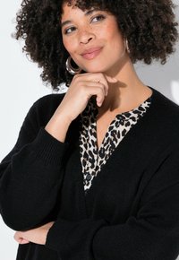 Zwarte gebreide cardigan met een diepe V-hals, gedragen over een blouse met luipaardprint. Opvallende kenmerken zijn de knoopdetails en de geribde manchetten.