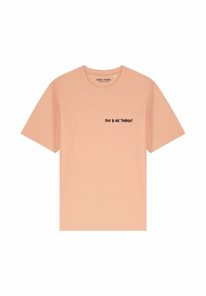 Jonge vrouw met lang blond haar die een perzikkleurig shirt draagt met de tekst "YOU & ME TONIGHT", staand in een park met historische gebouwen en een reuzenrad.