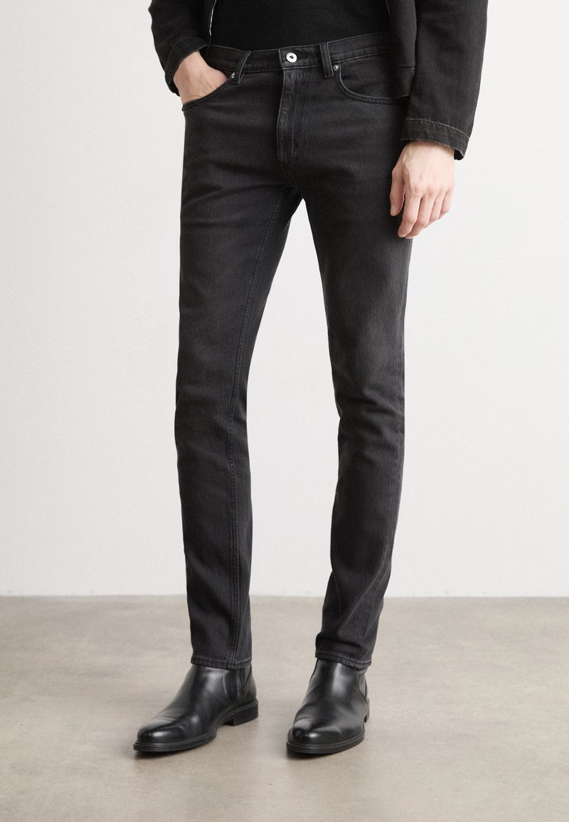 Black slim-fit jeans made of denim feature a mid-rise waist, five-pocket design, and subtle stitching. Paired with black ankle boots.