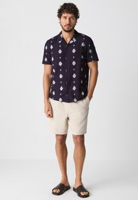 Kortærmet mørkeblå skjorte med hvid ikat mønster, knap-ned design, parret med lys beige shorts og brune slip-on sandaler.