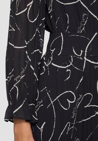 Schwarzes Kleid mit weißen Herz- und Schriftmustern. Verfügt über lange, plissierte Ärmel mit elastischen Bündchen und einer taillierten Passform. Leichtes Material.