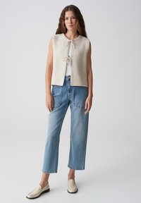 Gilet a maglia color crema con chiusura frontale a lacci, abbinato a jeans azzurri a vita alta e mocassini beige. Design semplice e moderno.