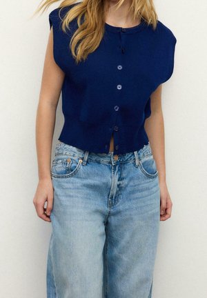 Kvinde iført en ærmeløs marineblå knap-oppet sweater og lyseblå højttaljede vidbenede jeans mod en ensfarvet baggrund.