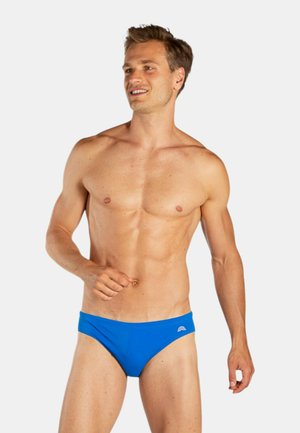 Uomo sorridente con i capelli corti in piedi, indossa slip da bagno blu acceso e posa con sicurezza contro uno sfondo chiaro e semplice.