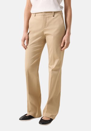 Kvinde iført beige bukser med lige ben og sorte balletsko, stående med afslappede hænder langs siden mod en lys baggrund.