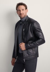 Czarna skórzana kurtka z wysokim kołnierzem, pikowanymi ramionami, zamkiem błyskawicznym z przodu i kieszeniami. Nosi się ją na szarym golfie.