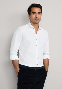 Witte overhemd met lange mouwen, opgerolde manchetten en kleine bruine knopen. Draagt met donkere broek en een marineblauwe riem. Soepele stoftextuur.
