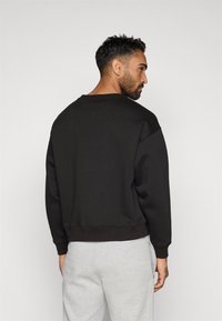 Sweatshirt preto com decote redondo e mangas longas, apresentando punhos e bainha canelados. Combinado com calças de moletom cinzentas, visto por trás.