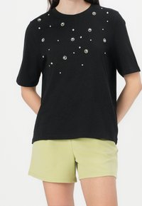 T-shirt noir à manches courtes ornée de détails blancs et argentés dispersés sur le devant, assorti à un short vert clair.