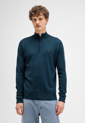 Man with curly hair wearing a textured dark teal half-zip sweater and light blue jeans, standing against a plain light background.