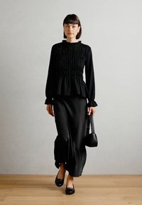 Černé dlouhé tričko s texturovaným smockingem, spárované s volnou černou sukní. Matné černé baleríny a malá kabelka přes rameno dotvářejí celý outfit.