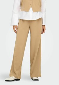 Pantalons larges beiges en tissu doux ; dotés d'une texture lisse avec une coupe décontractée ; associés à une chemise blanche à boutons et des chaussures claires.