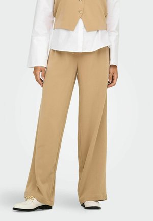 Pantalons larges beiges en tissu doux ; dotés d'une texture lisse avec une coupe décontractée ; associés à une chemise blanche à boutons et des chaussures claires.