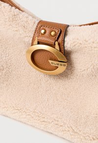 Beżowa torba na ramię z owczej skóry, z brązowym skórzanym paskiem, z okrągłą złotą klamrą z wygrawerowanym napisem "GUESS". Teksturowane i miękkie materiały.