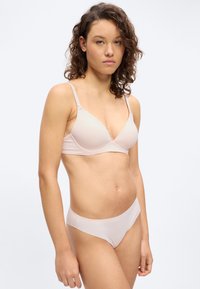 Νεαρή γυναίκα με σγουρά μαλλιά, που φοράει ασορτί ροζ ανοιχτό σουτιέν και εσώρουχα, στέκεται μπροστά σε απλό φωτεινό φόντο, στραμμένη ελαφρώς προς τα αριστερά.