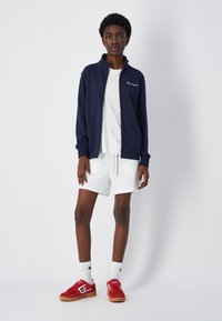 Sudadera azul marino con cremallera y cuello alto, logo blanco, llevada sobre una camiseta blanca y pantalones cortos blancos; zapatillas rojas con detalles blancos.