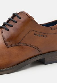 Chaussure habillée en cuir marron à texture lisse, avec un bout arrondi et des lacets noirs. Présente des coutures discrètes et un logo « bugatti » embossé.