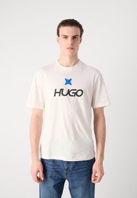 Cream-colored cotton t-shirt with short sleeves, featuring a black "HUGO" logo and a blue star accent in the center. Casual fit and simple design.
