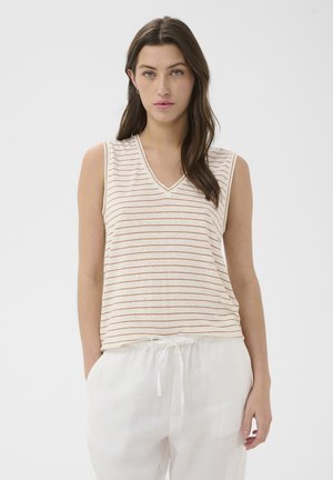 Femme aux longs cheveux bruns portant un haut beige sans manches à fines rayures rouges et un pantalon blanc ample à cordon, debout devant un fond blanc.