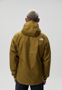 Khaki vodeodolná bunda s kapucňou, elastickými manžetami a logom na zadnej strane. Hladká, lesklá textúra; navrhnutá na vonkajšie použitie.