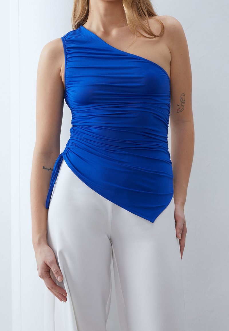 Femme portant un haut bleu froncé à une épaule avec un ourlet asymétrique, associé à un pantalon blanc à jambes larges, tatouages visibles sur les bras.