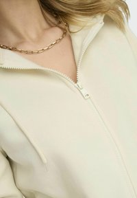 Felpa con zip color crema, caratterizzata da una cerniera frontale con un tirante testurizzato e dei lacci. Accompagnata da una collana a catena dorata.