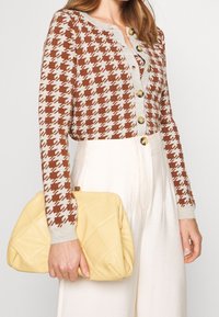 Femme portant un cardigan pied-de-poule marron et beige avec des boutons dorés, un pantalon blanc taille haute, tenant une pochette matelassée jaune pâle.