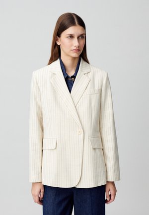 Blazer bianco sporco con sottili righe verticali, chiusura a un bottone, rever a punta e due tasche frontali. Indossato sopra una camicia di jeans.