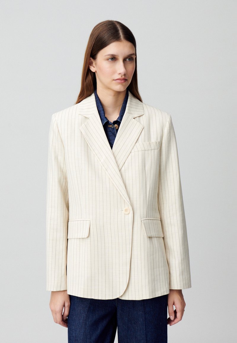 Blazer bianco sporco con sottili righe verticali, chiusura a un bottone, rever a punta e due tasche frontali. Indossato sopra una camicia di jeans.