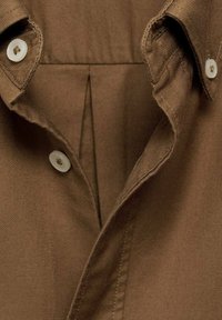 Brown button-up shirt featuring a collar, two white buttons, and a clean, textured fabric. Includes a back pleat for ease of movement.