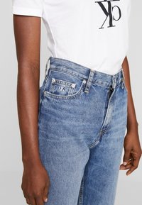 Person som bär ljusblå Calvin Klein-jeans med en vit logotröja, synliga midje- och övre lårområden, händerna avslappnat längs sidorna.