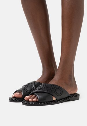 Sorte lædersandaler med krydsede stropper, tekstureret overflade, dekorative nitter og en flad flettet sål. Åben tå-design.