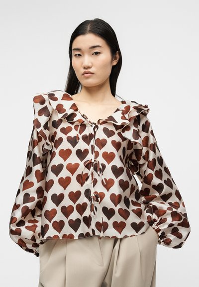 Γυναίκα που φοράει μπλούζα με καφέ σχέδια καρδιάς, βολάν γιακά, μακριά φουσκωτά μανίκια και μπεζ παντελόνια μπροστά σε απλό φωτεινό φόντο.
