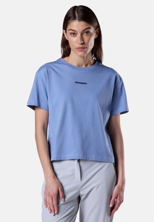 Kobieta o prostych brązowych włosach ubrana w luźną, jasnoniebieską koszulkę Rossignol i jasnoszare spodnie, stoi na tle jednolitego, jasnego tła.