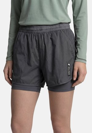 Personne portant un short de sport superposé gris foncé et un haut à manches longues vert clair, debout les bras relâchés de chaque côté.