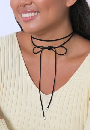 Black choker necklace made of cord with a bow detail, featuring two silver metal ends. Worn with a textured light yellow top.