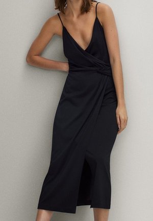 Donna che indossa un vestito midi nero senza maniche con scollo a V profondo e dettaglio incrociato, in piedi contro un muro neutro, mano appoggiata sul fianco.