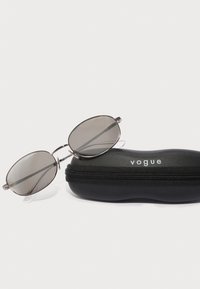 Oválne sivé slnečné okuliare so striebornými kovovými rámami, odložené na čiernom zipsovom púzdre, ktoré nesie značku "vogue."