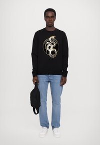 Felpa nera con grafiche di un serpente in oro, abbinata a jeans blu chiaro e scarpe da ginnastica bianche. Il modello tiene una borsa nera.