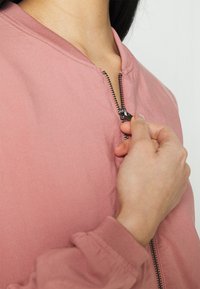 Giacca bomber rosa chiaro con chiusura a zip, colletto a coste e maniche a costine. Il tessuto appare liscio e leggermente testurizzato.