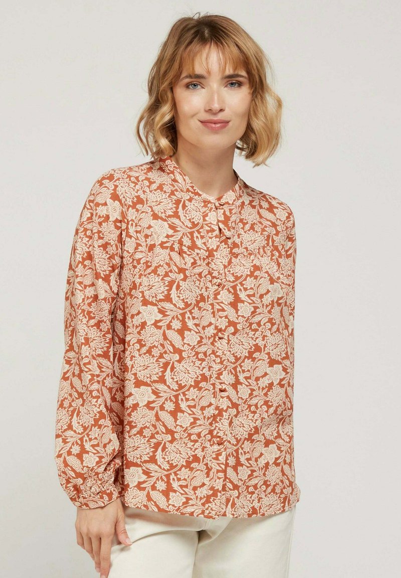 Blouse à manches longues de couleur rouille avec un motif floral blanc, fermeture à boutons sur le devant, col rond et poignets froncés. Tissu léger.